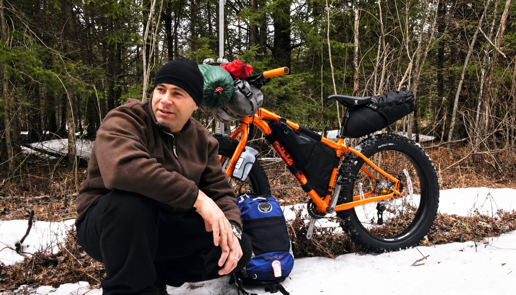 Bikepacker resting beside a loaded orange fat bike in a snowy forest